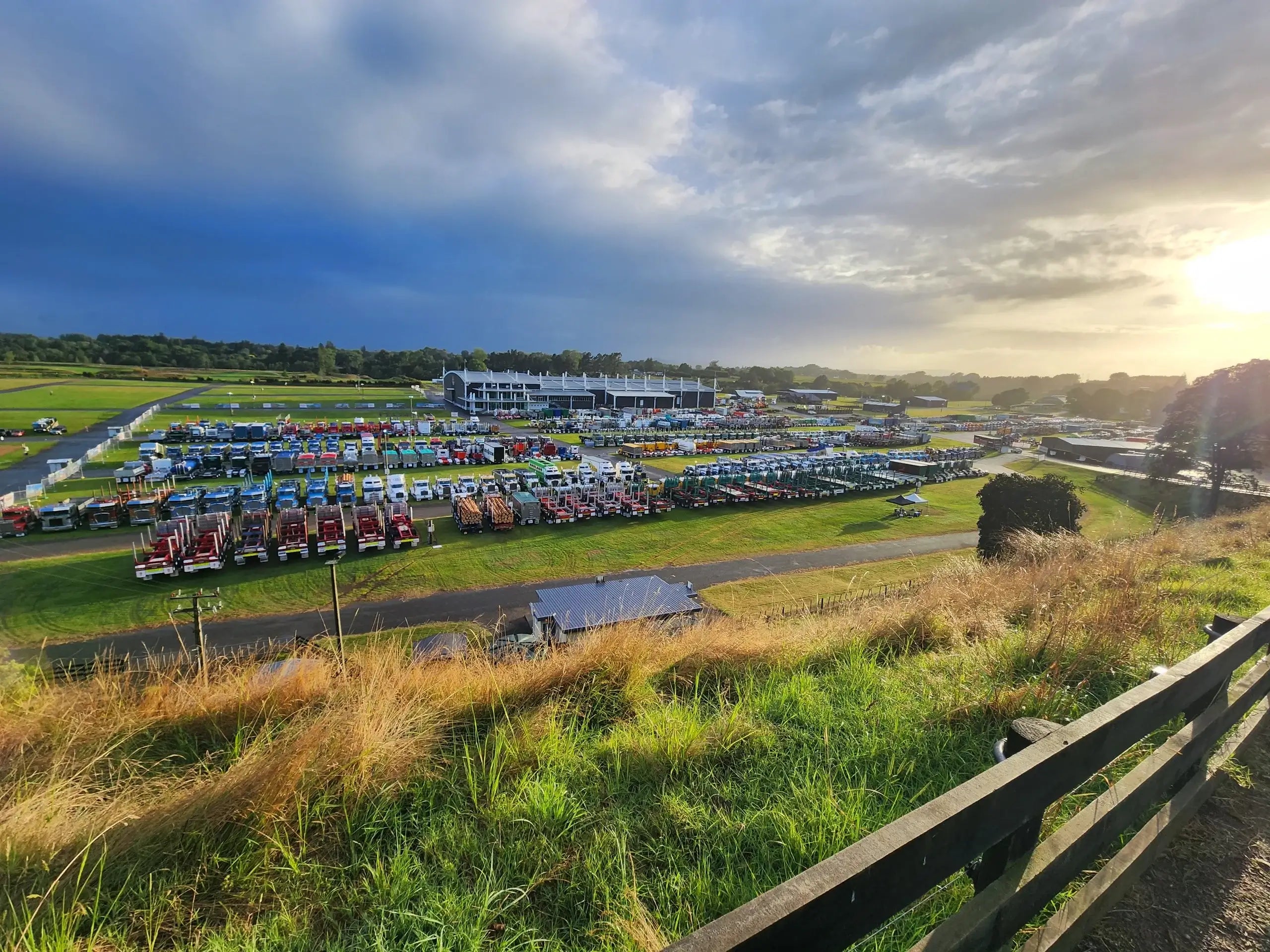 A panoramic view of a large outdoor event with rows of colorful trucks parked on a grassy field, a building in the background, and the sun setting behind clouds, casting a golden light over the scene.