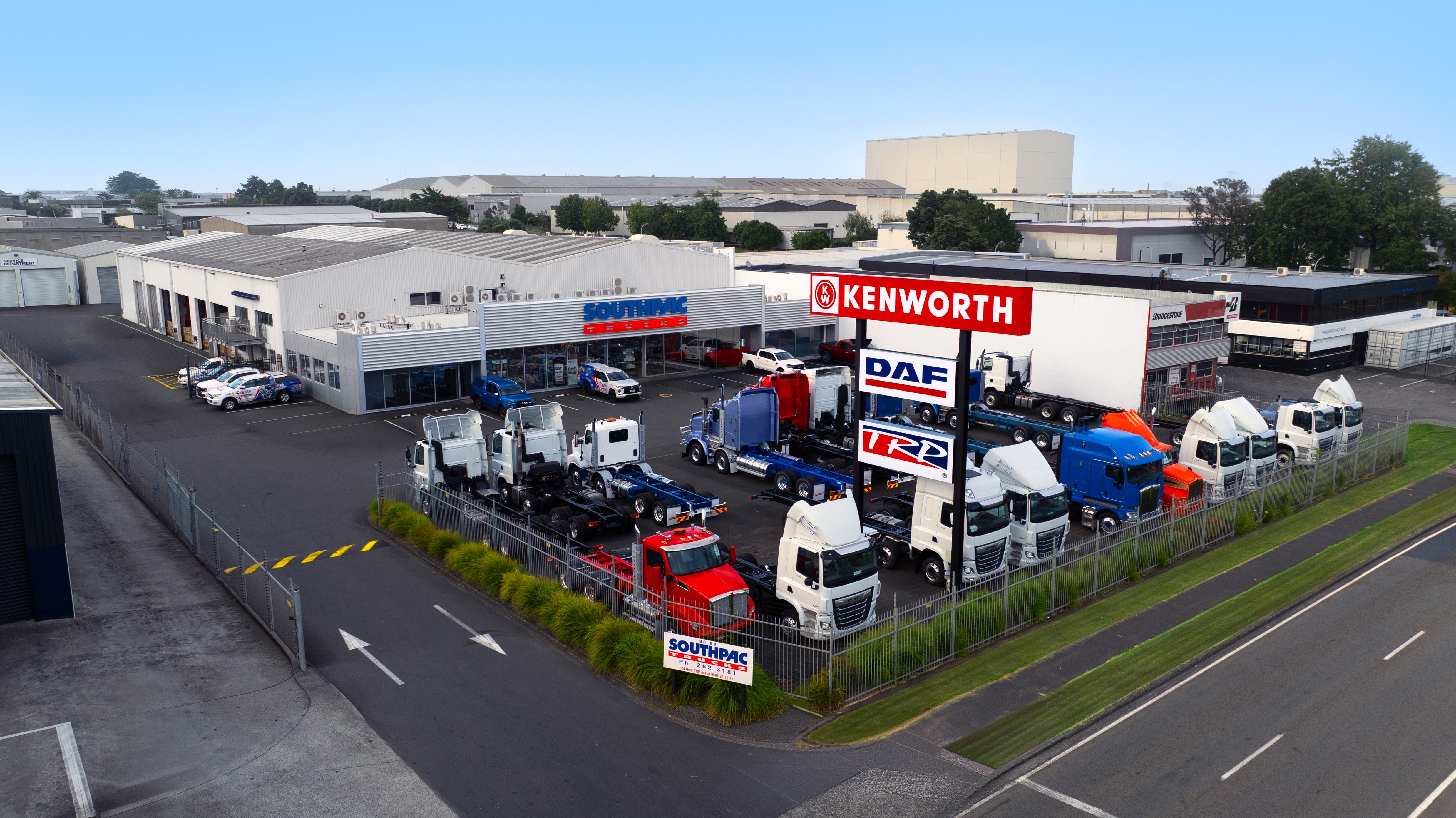 A fenced truck dealership displays multiple Kenworth, DAF, and other trucks next to a large building, with signs and parked cars visible. The area is industrial, with warehouses in the background.
