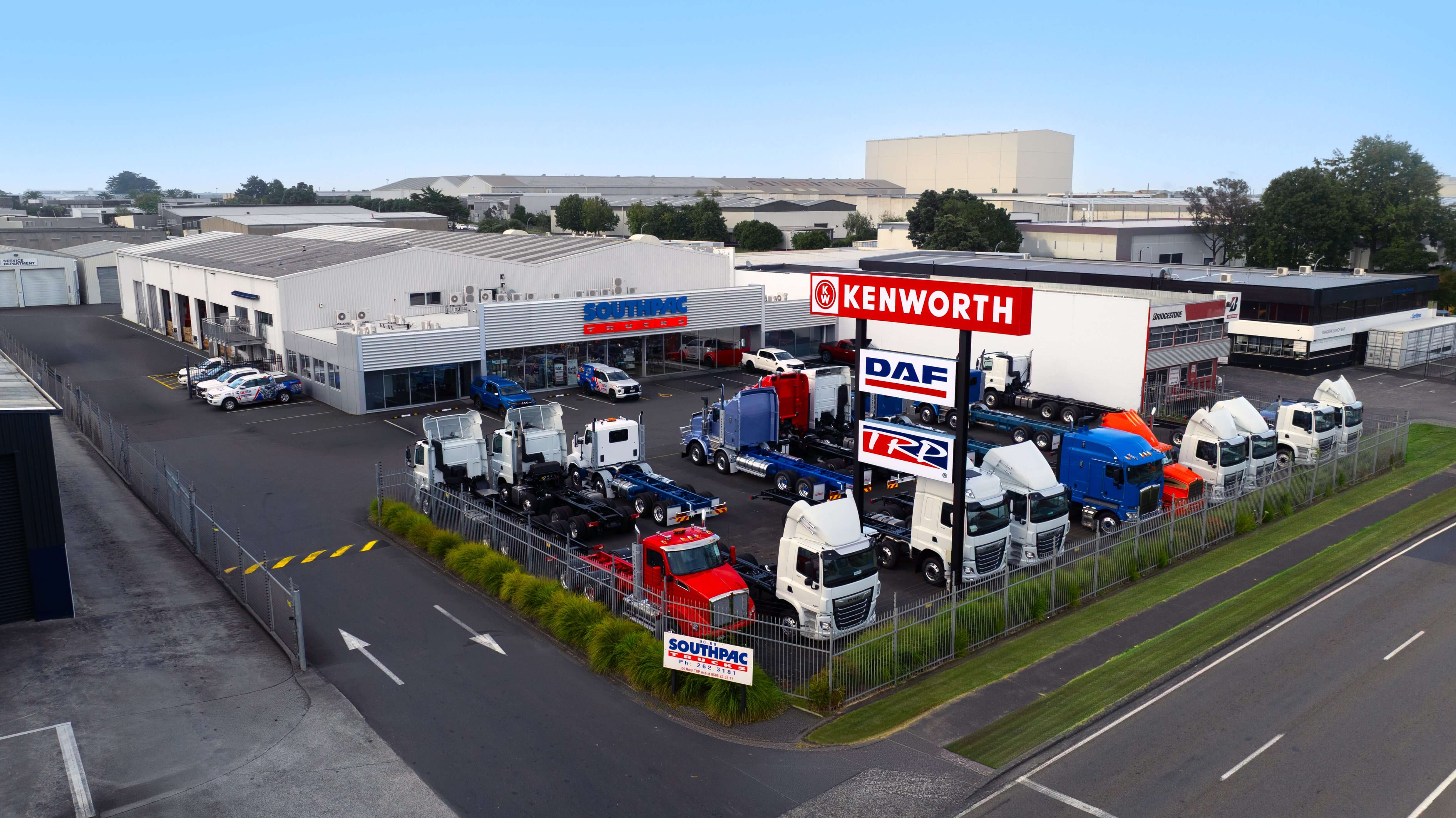 A fenced truck dealership displays multiple Kenworth, DAF, and other trucks next to a large building, with signs and parked cars visible. The area is industrial, with warehouses in the background.