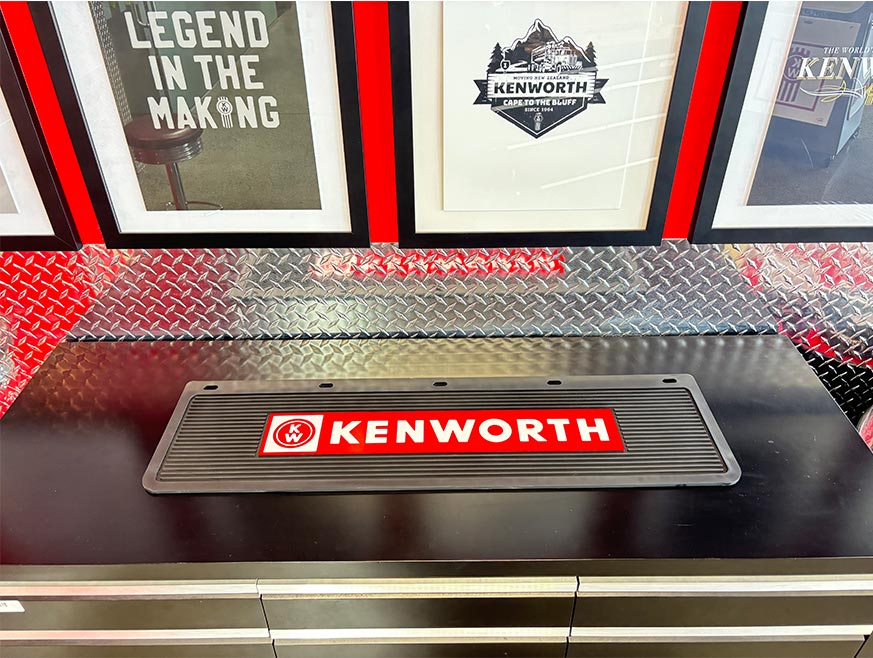 The BAR MAT KW RUBBER sits on a black tool chest, protecting the countertop, while framed Kenworth posters and diamond plate metal decor complete the look.
