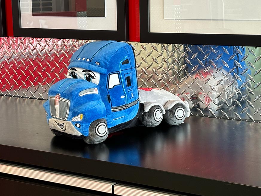 TOY PLUSH KATIE, a soft and durable blue semi-truck with cartoon eyes and a smiling face, sits on a black countertop before a diamond plate wall—making it the perfect gift for kids.
