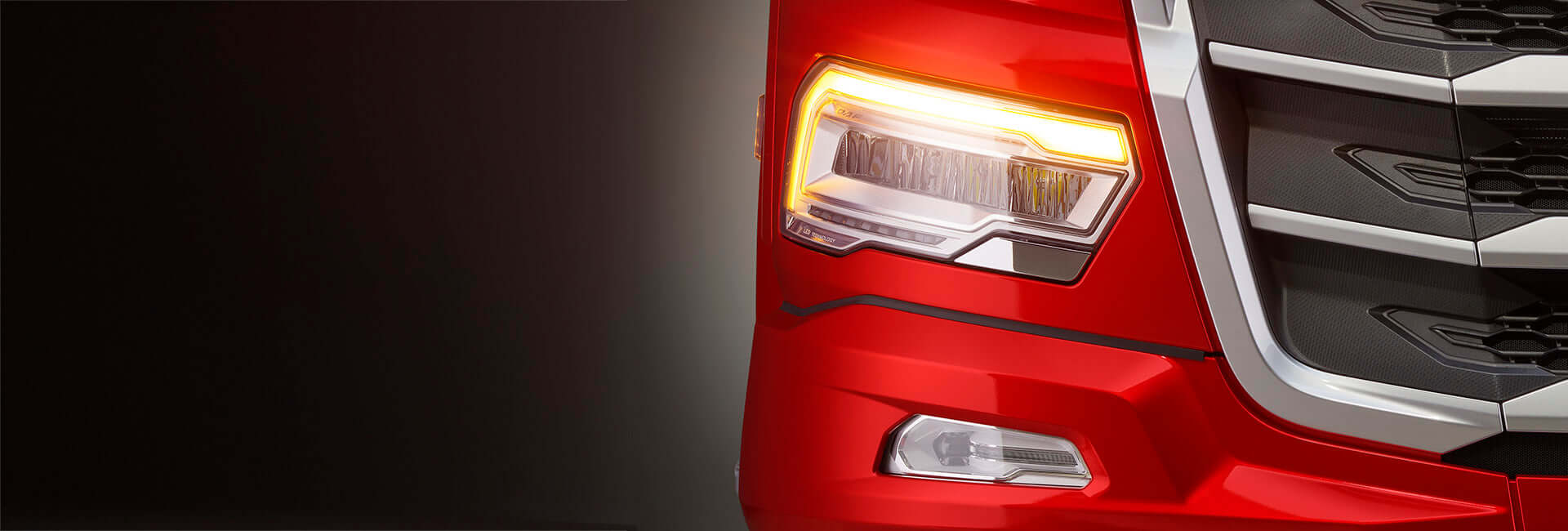 Close-up of the front right corner of a red vehicle, showing a bright amber headlight and part of the chrome grille against a dark background.