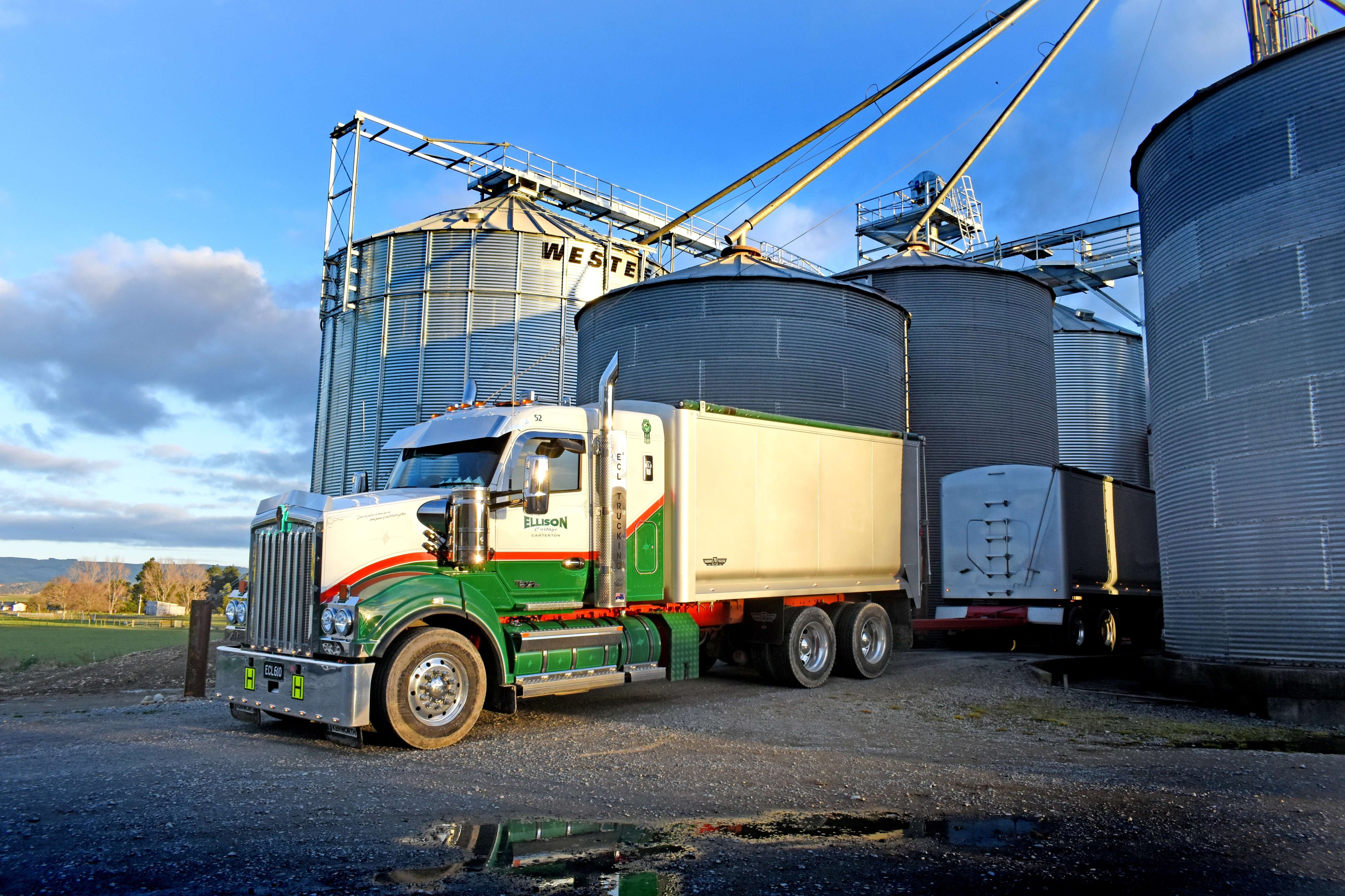 A large green and white semi-truck is parked next to metal grain silos at a farm, with blue sky and machinery visible in the background. The truck is positioned on a gravel area near the silos.