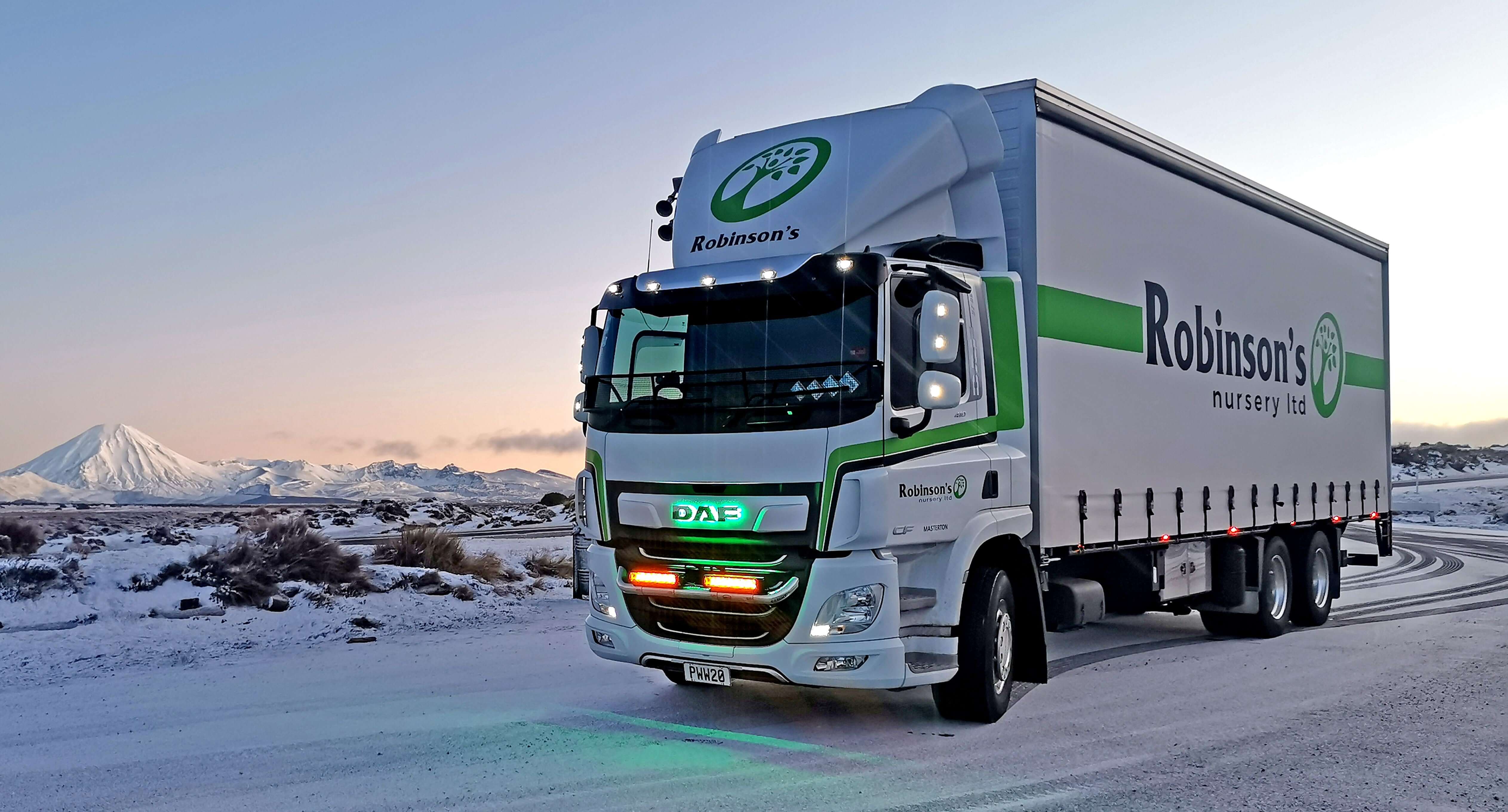 A Robinson’s Nursery Ltd truck drives on a snowy road with snow-covered mountains in the background during sunset or sunrise.