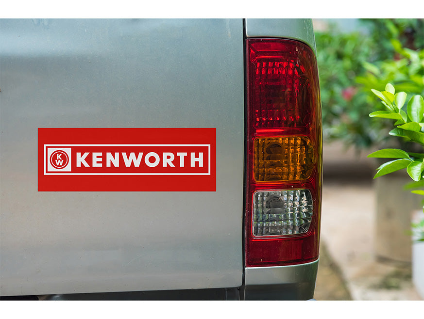 Close-up of a gray vehicle's rear with a red and white KENWORTH Banner Bumper Sticker by the taillight. Green plants are visible in the background on the right.