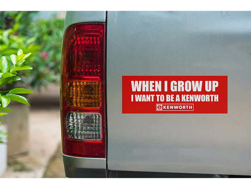 A gray vehicle displays the KENWORTH When I Grow Up Sticker—a red decal reading "WHEN I GROW UP I WANT TO BE A KENWORTH" in white text with the Kenworth logo below. A tail light and green plants appear in the background.