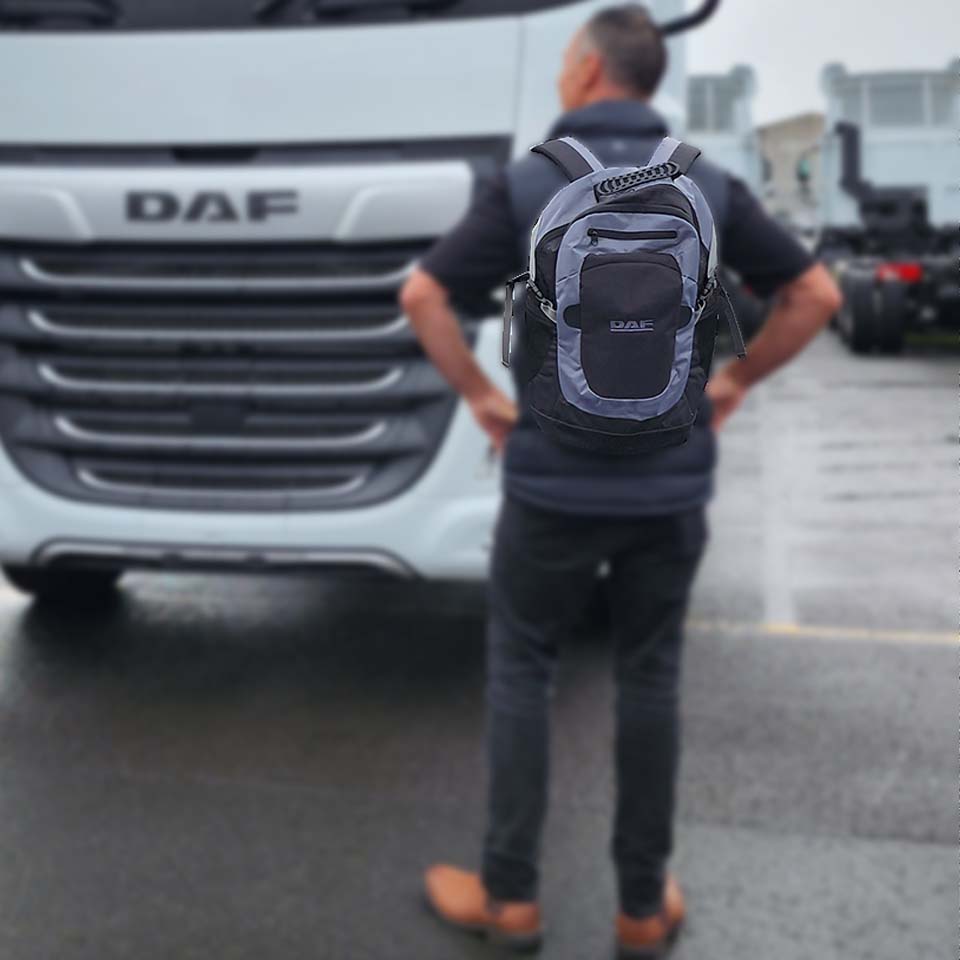 A person with the gray and black BACKPACK DAF stands on wet pavement, facing a white DAF truck outdoors, possibly in a parking or industrial area.