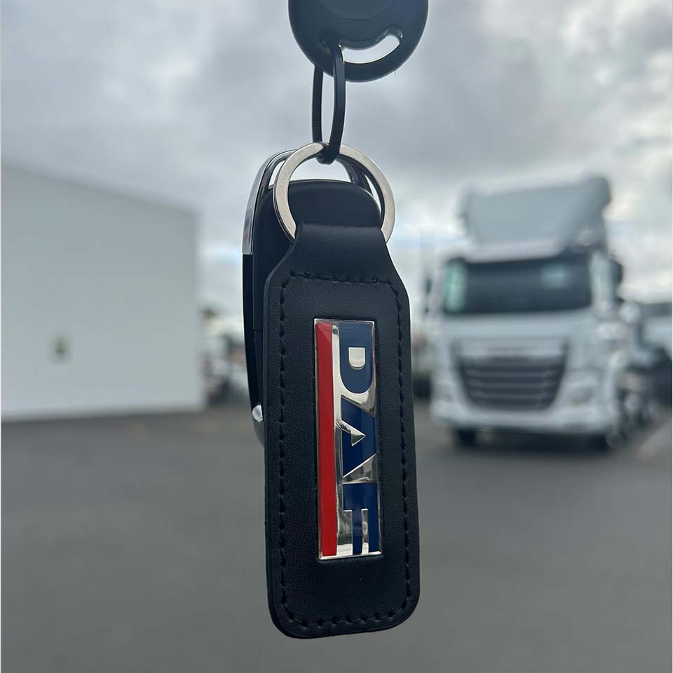 A close-up of the KEYRING DAF LEATHER shows the DAF logo in silver, red, and blue on black leather. It hangs outdoors with a white truck and building in the blurred background—an ideal accessory for DAF fans.