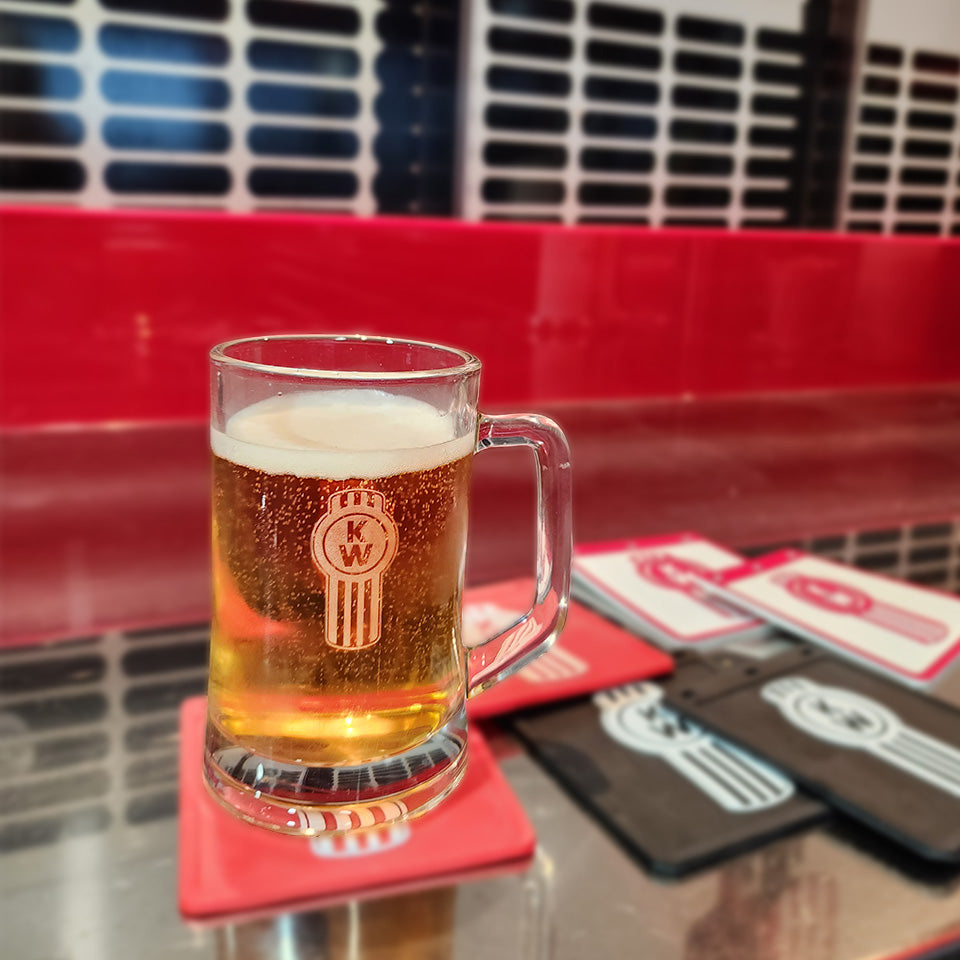 The GLASS KW BEER HANDLE, filled with frothy beer, rests on a red coaster atop a shiny counter. Several coasters and a red wall with graphic design in the background create an inviting home bar vibe.