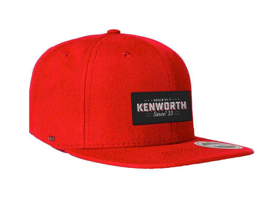 The CAP KW HAULIN 24-7 RED is a bright red cap with a structured crown and flat brim, featuring a black patch on the front that says "Kenworth Since 23" in white. The snapback closure offers adjustable sizing for the perfect fit.