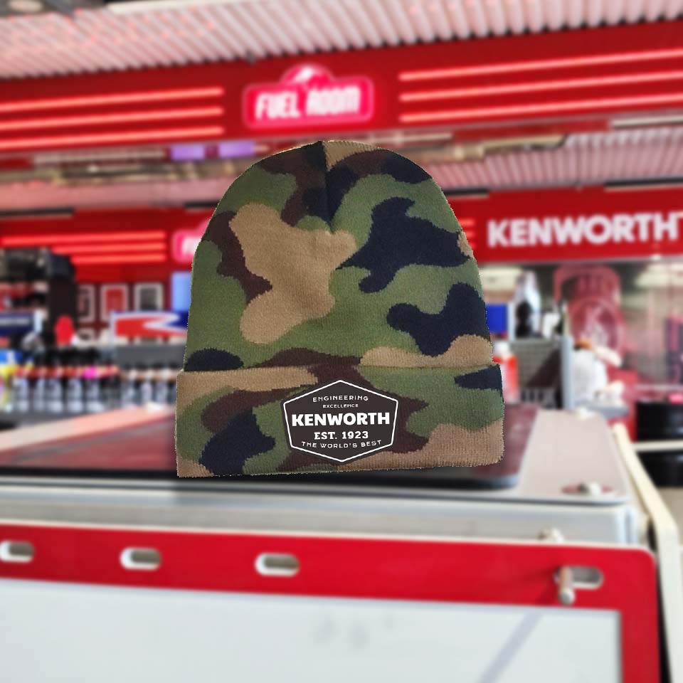 The BEANIE KW EXCELLENCE CAMO, featuring a KENWORTH EST. 1923 patch, is displayed in focus with a blurred convenience store and a bold red KENWORTH sign in the background, highlighting Kenworth Engineering Excellence.
