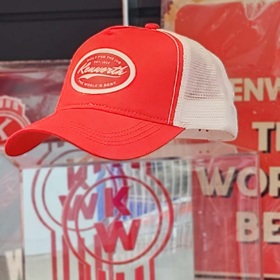 The CAP KW BUILT FOR THE JOB RED, a red and white Kenworth trucker hat with mesh back, is shown indoors against blurred Kenworth logos and red signage, showcasing The World's Best classic trucking style.
