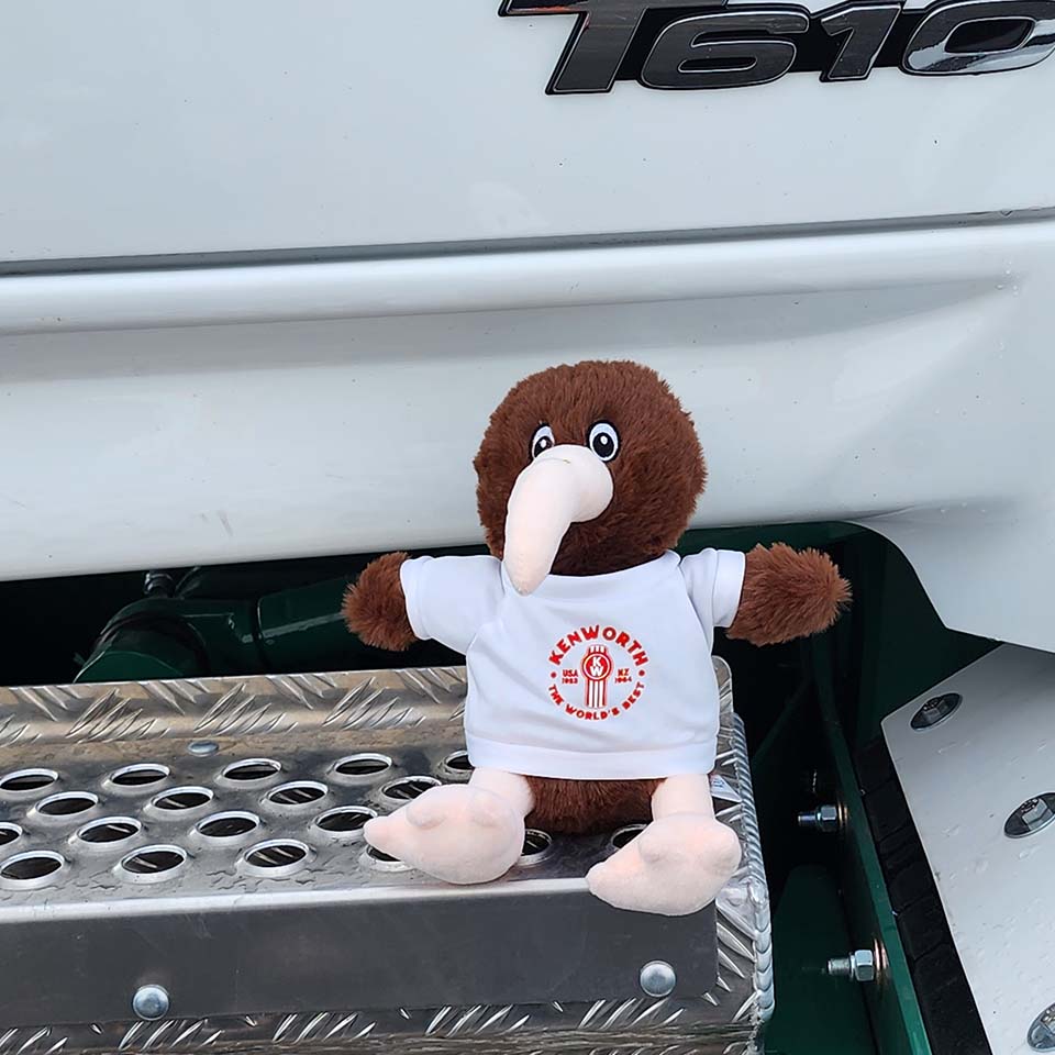 TOY KW KEV THE KIWI SOFT TOY, sporting a white Kenworth tee, sits on a truck step next to a white T610—an ideal keepsake for Kenworth truck enthusiasts.