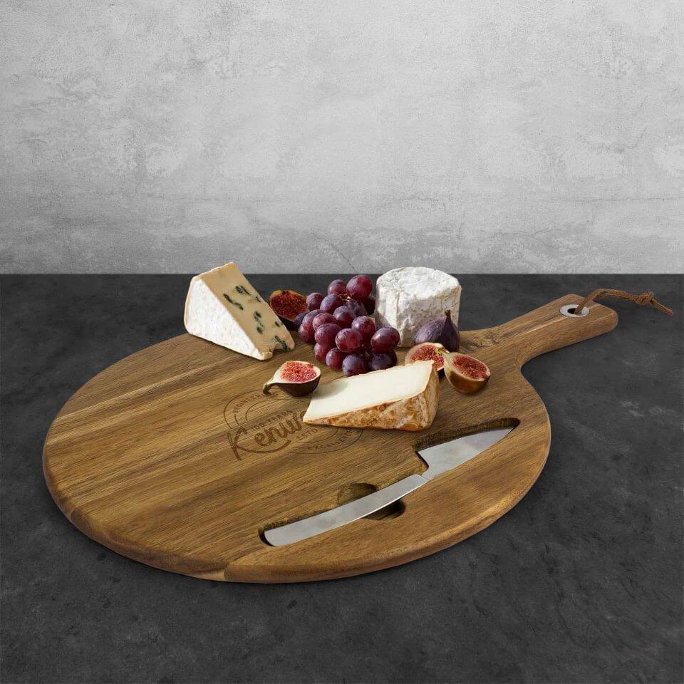 The WOOD BOARD AND KNIFE SET, arranged with three cheeses, red grapes, and halved figs, is displayed on a dark countertop against a light gray textured wall.