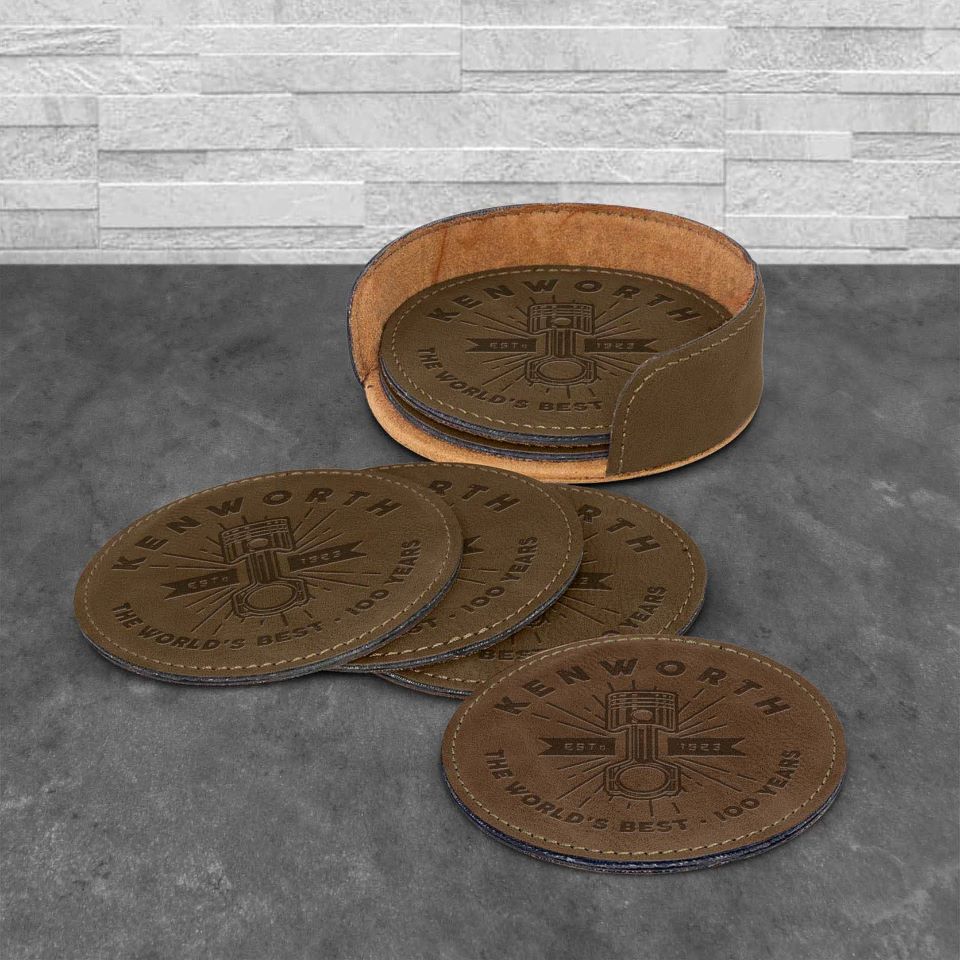 The COASTER KW BROWN set includes five round brown PU coasters featuring the Kenworth The Worlds Best 100 Years 1923-2023 logo. Four coasters are arranged on a gray surface and one is placed in the matching holder.