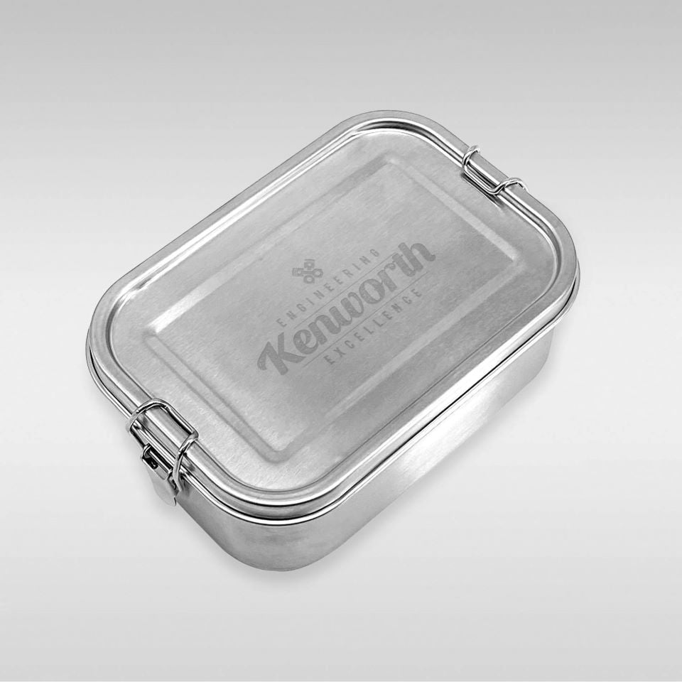 The LUNCH BOX KW S/S is a rectangular stainless steel lunch box with rounded edges and two side clasps. "Engineering Kenworth Excellence" is engraved on the lid. The background is plain, light grey.