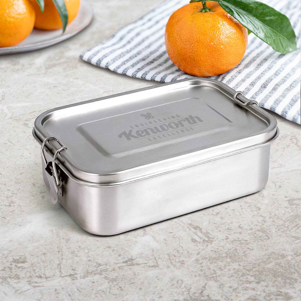 A food-grade, stainless steel LUNCH BOX KW S/S with side clasps rests on a light countertop near a striped cloth and an orange. The eco-friendly lunch box is ideal for everyday use.