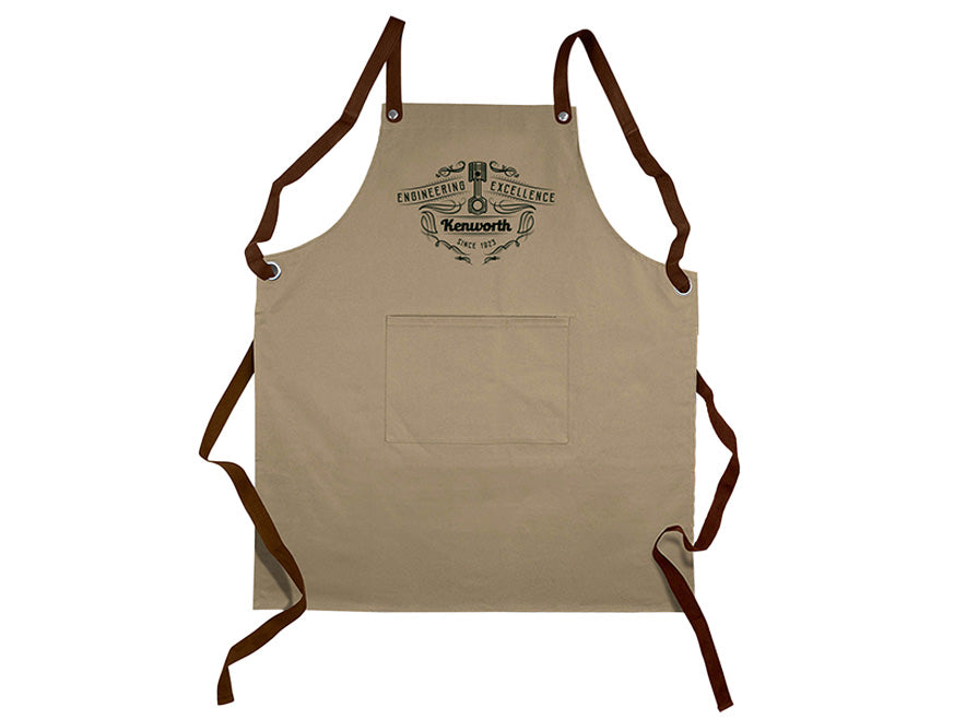 The APRON KW TAUPE is a khaki cotton bib apron with brown adjustable cross-back straps, a front pocket, and the Kenworth logo plus "Engineering Excellence" and "The World’s Best" printed above the pocket in black.