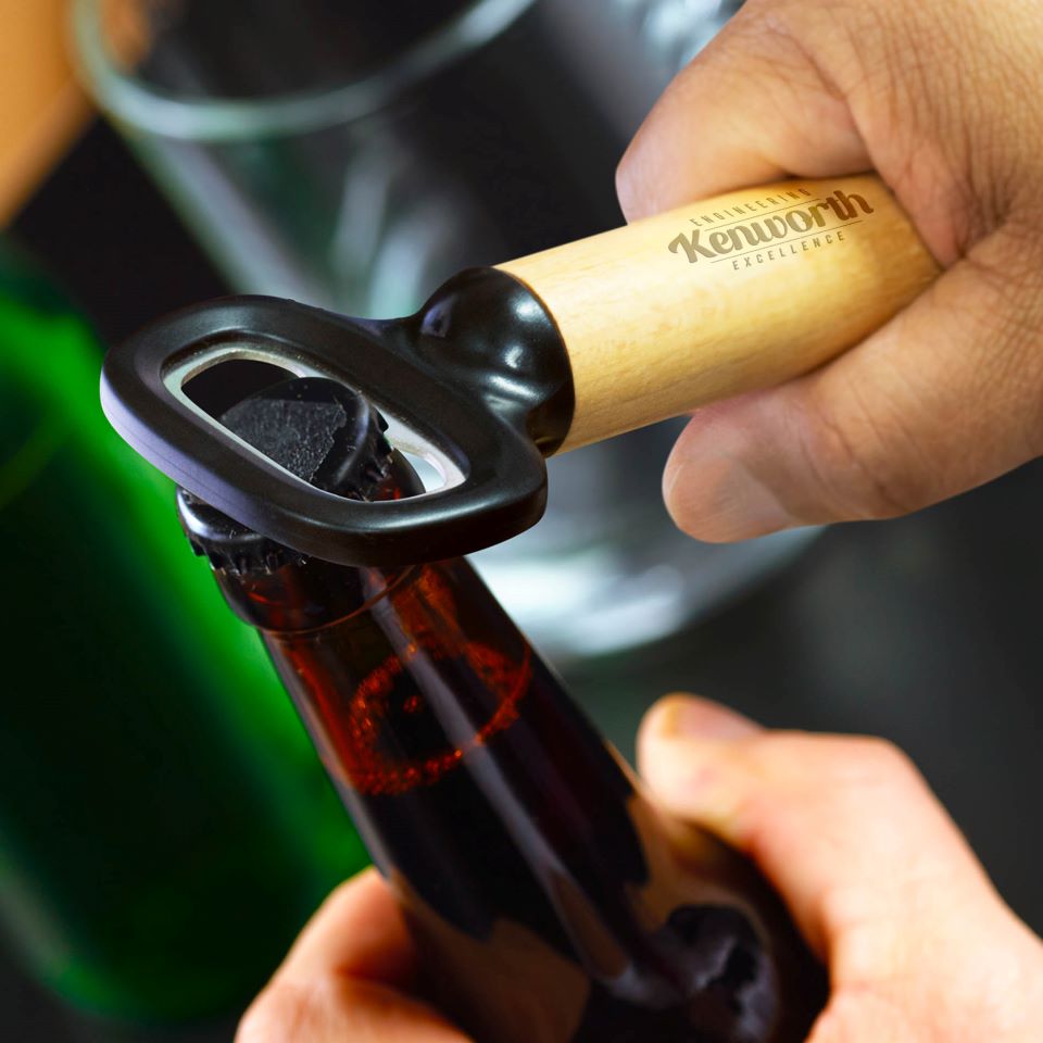 A hand uses the BOTTLE OPENER KW WOOD & S/S to open a brown glass bottle, with a green bottle and a glass blurred in the background.