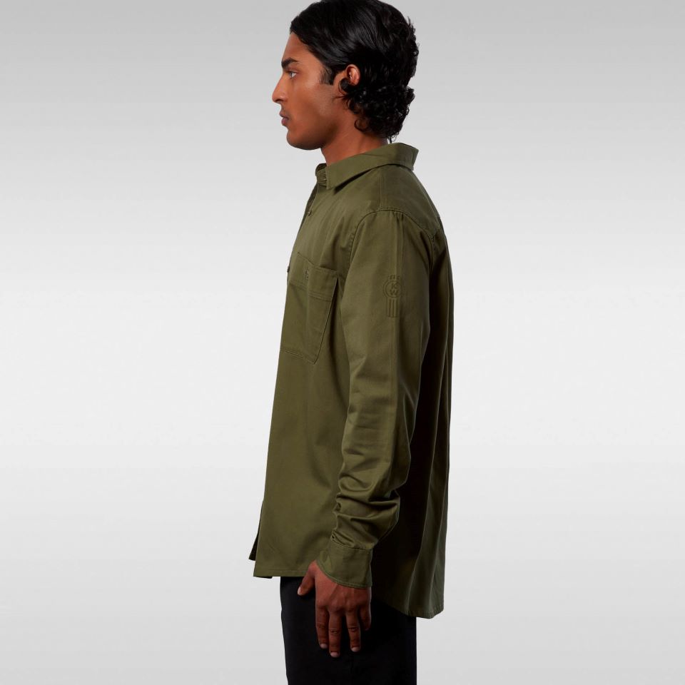 A man with dark, wavy hair stands in profile against a plain light gray background, wearing the SHIRT KW DRILL ARMY in olive green and black pants—an ideal choice for professional wear.