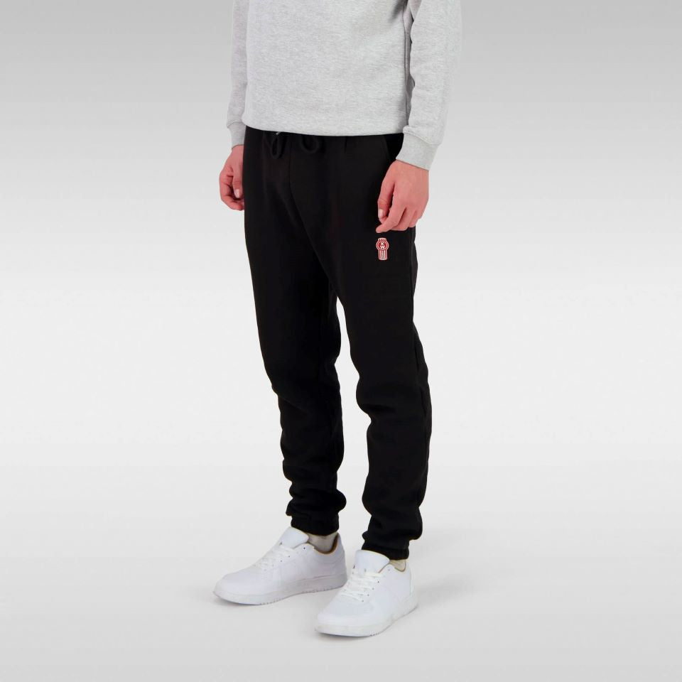 Model wearing SWEATPANT KW MENS BLACK with a small red logo on the left thigh, paired with a light gray sweatshirt and white sneakers, standing against a plain light gray background. Only the lower half is visible.
