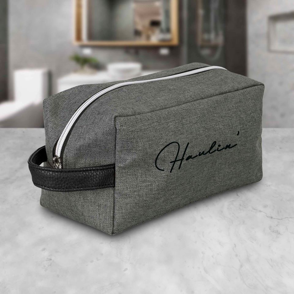 The BAG KW TOILETRY is a gray rectangular travel toiletry bag with a strong handle and white zipper, featuring "Hauline" in black cursive on the side. It rests on a marble surface with a blurred bathroom in the background.