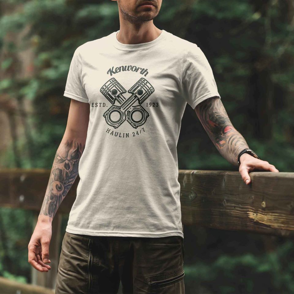 A person with tattooed arms wears the TEE SHIRT KW HAULIN SAND, featuring "Estd. 1923 Haulin’ 24/7" and crossed pistons, standing outdoors by a wooden railing with trees in the background.