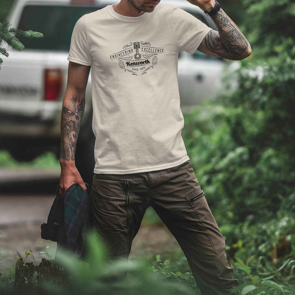 A person with tattooed arms wears a TEE SHIRT KW EXCELLENCE and khaki cargo pants, standing outdoors in a green area near a parked vehicle, holding a jacket in one hand.