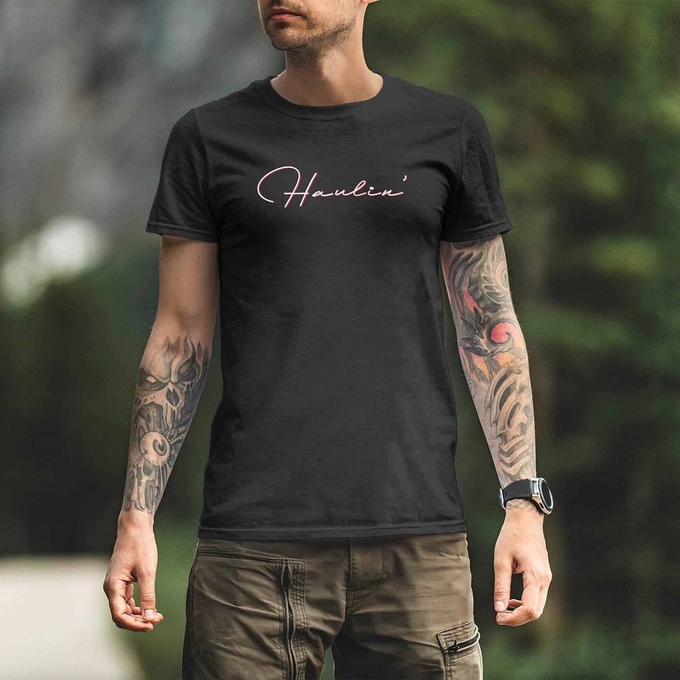 A tattooed man wears the TEE SHIRT KW HAULIN BLACK, featuring a cursive Howlin' design. He stands outside, with blurred greenery and trees in the background.
