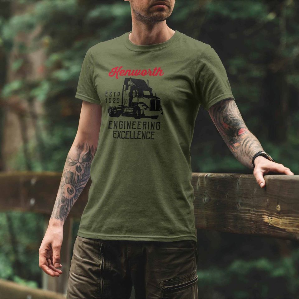 A tattooed man wears a TEE SHIRT EXCELLENCE GREEN with a Kenworth truck graphic, standing outdoors on a wooden bridge surrounded by greenery.