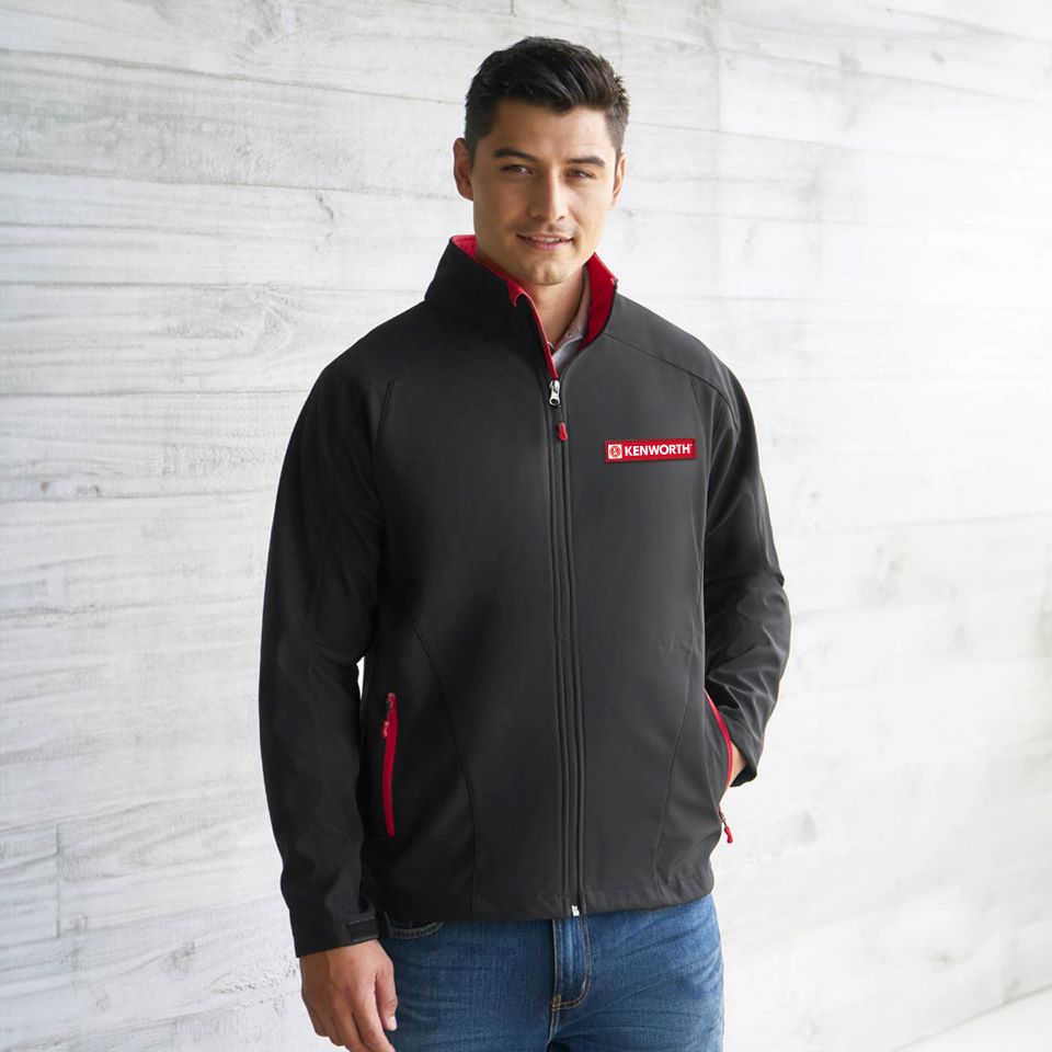 A man stands against a light gray wall wearing the JACKET KW GENEVA, which is black with red accents. He pairs it with blue jeans, has short dark hair, smiles slightly, and poses with one hand in his pocket for a relaxed vibe.