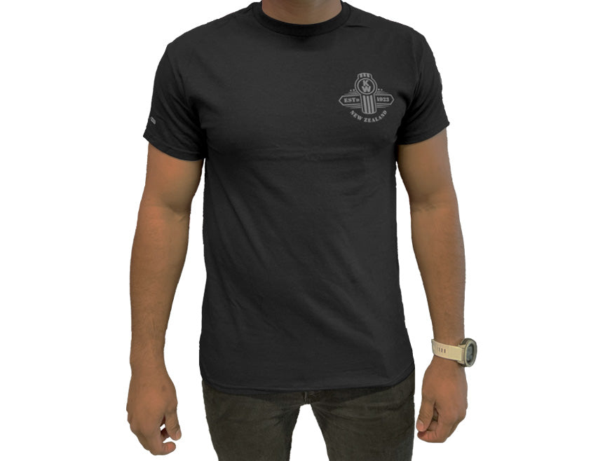 Someone wearing the TEE SHIRT KW 100 YEARS—a plain black shirt with a small, circular white Kenworth 100 years logo on the left chest—pairs it with black pants and a light wristwatch, standing against a white background.