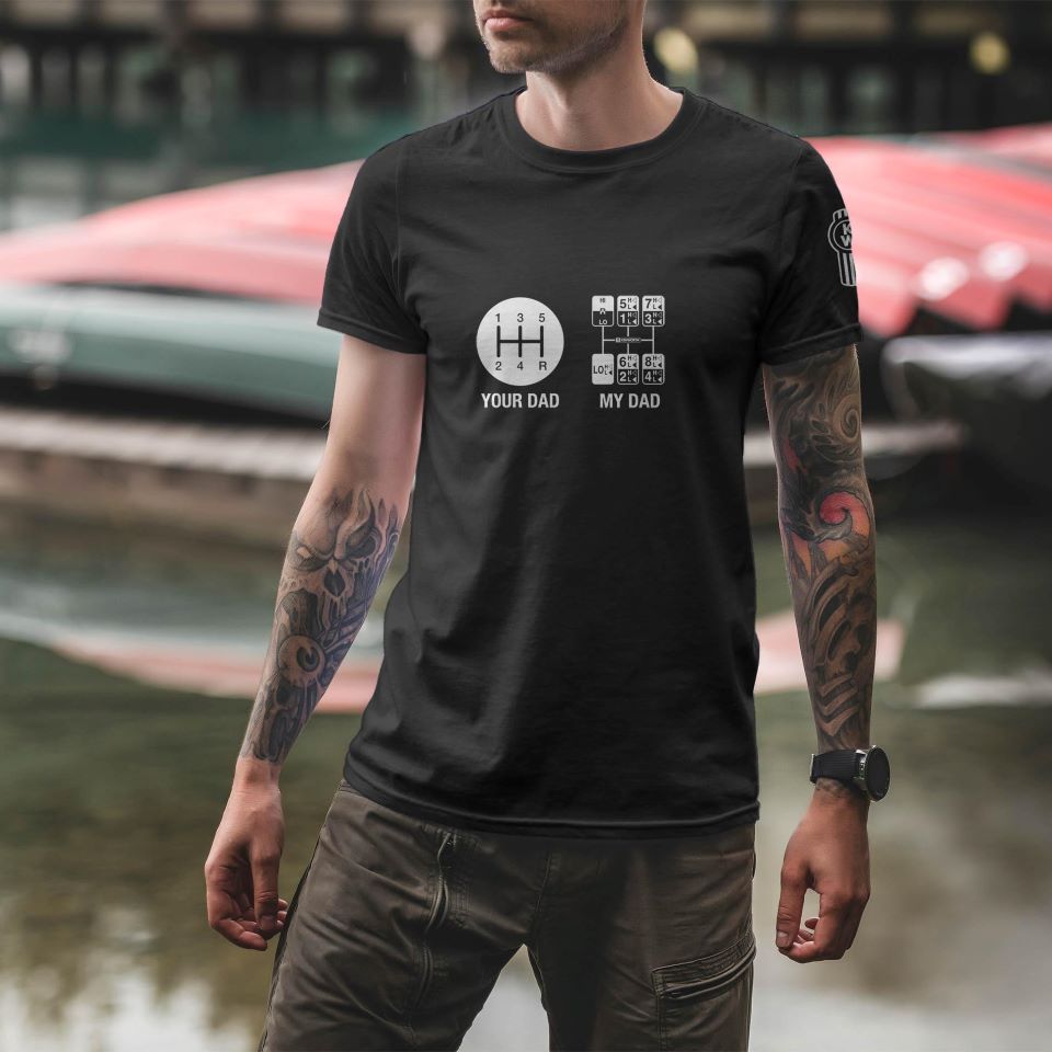 A tattooed man stands outdoors near boats, wearing the TEE SHIRT KW SHIFTER—a black graphic tee with "Your Dad My Dad Gear" shift diagrams.