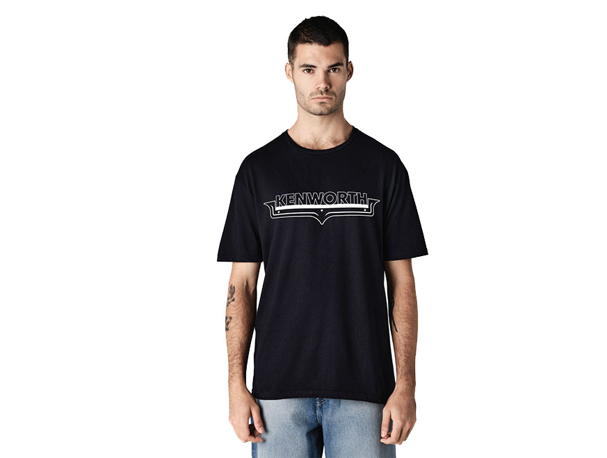 A man with short dark hair and light facial hair models the TEE SHIRT KW NAMEPLATE, which displays a bold Kenworth logo in white, paired with light blue jeans against a plain white background.