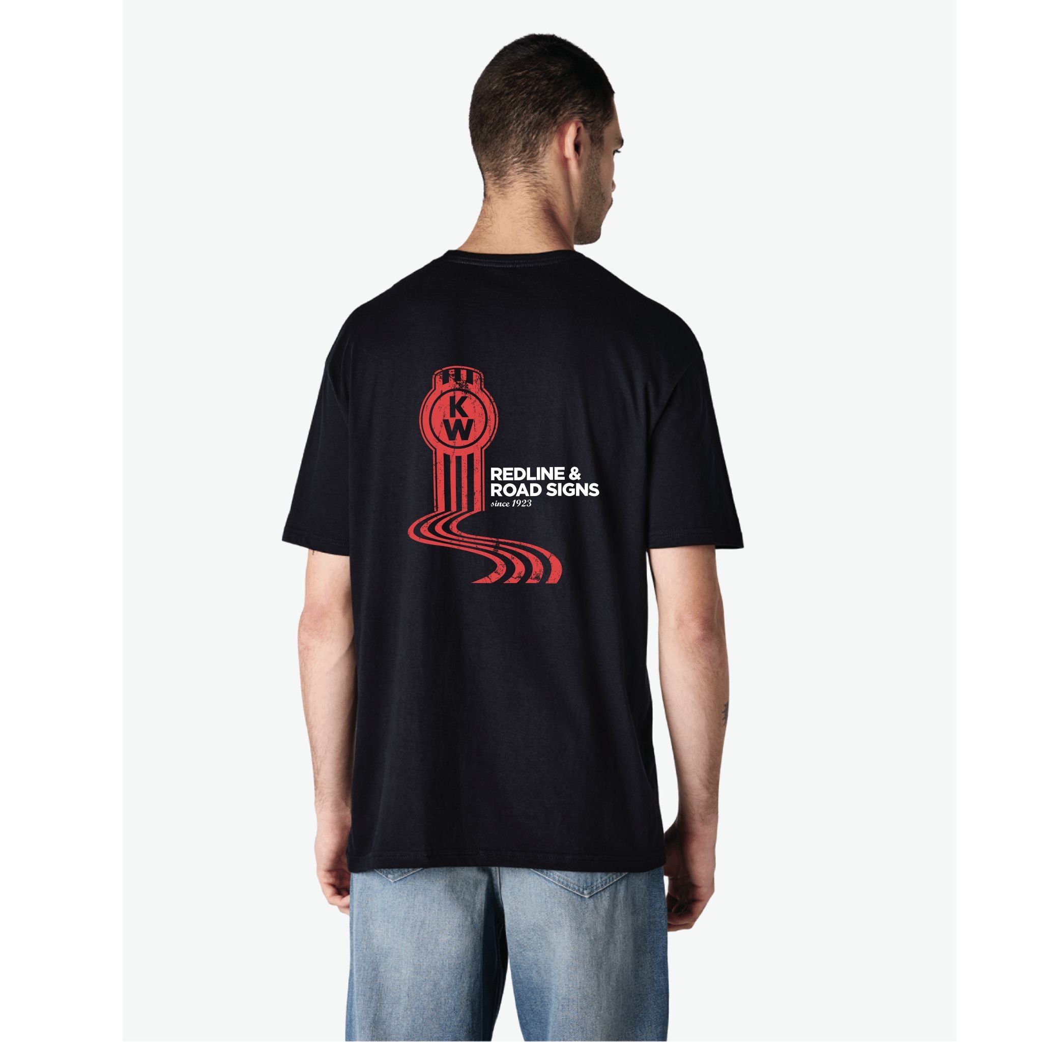A man wears the TEE SHIRT KW REDLINE 2XL, a black Kenworth tee with redline and road sign graphics plus a circular KW logo and "Redline & Road Signs (Part 2)" on the back, paired with light blue jeans.