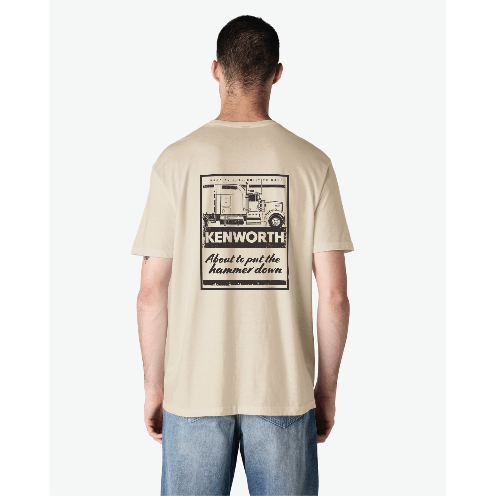 A person faces away from the camera wearing the TEE SHIRT KW HAMMER, a sand-colored Kenworth shirt with a bold semi-truck graphic and “Kenworth – About to put the hammer down” text on the back.