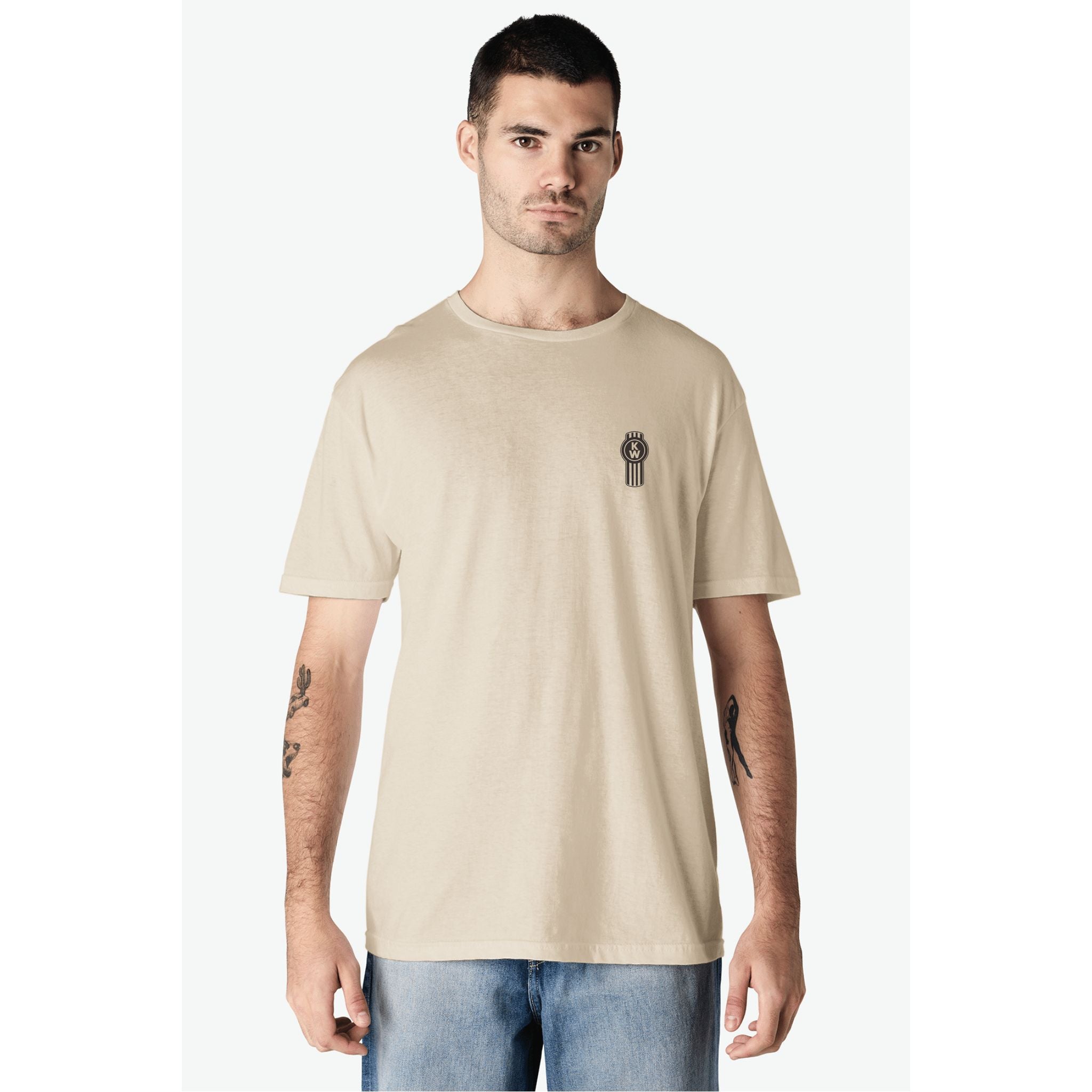 A man with short dark hair and tattooed forearms wears the TEE SHIRT KW HAMMER, a sand-colored tee with a small black and white chest graphic, paired with light blue jeans against a plain light background.