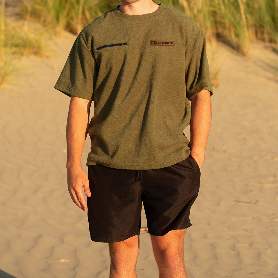 A person stands on sandy ground with tall grass behind, wearing the olive green SHIRT BUSH KW SHORT with a Kenworth patch on the chest and black shorts. Sunlight highlights this relaxed outdoor style.