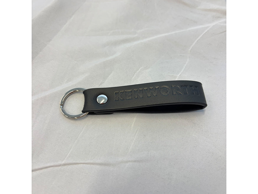 The KEYRING KW BLACK LOOP features black leather with a silver metal keyring and embossed Kenworth logo, shown on white fabric—a sleek accessory perfect for any Kenworth fan.