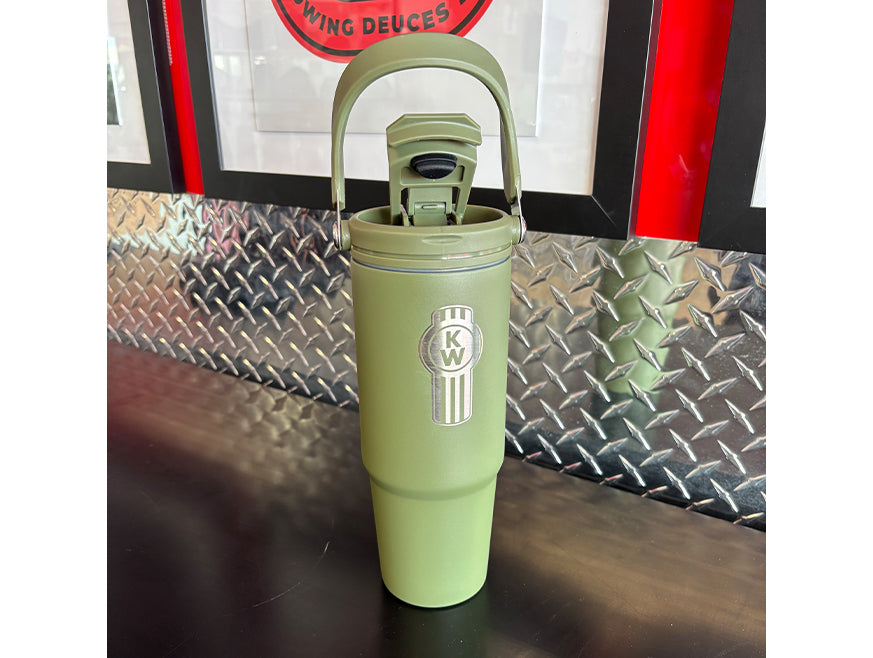 A green BOTTLE KW TRAVEL GREEN INSULAT with a handle and Kenworth logo sits on a black counter before a metal diamond plate wall and framed posters.