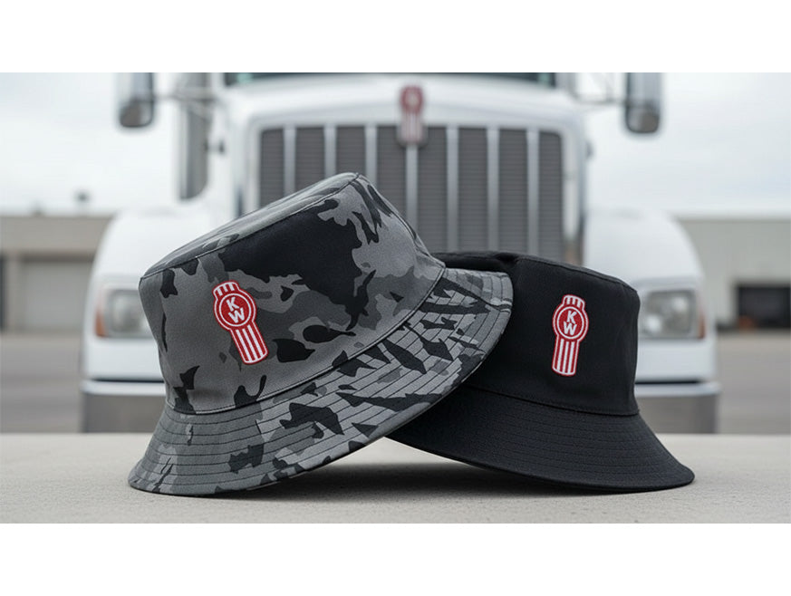 A Kenworth Bucket Black/Camo Reversible hat, featuring a red and white Kenworth logo, is shown in front of a white Kenworth truck grille.