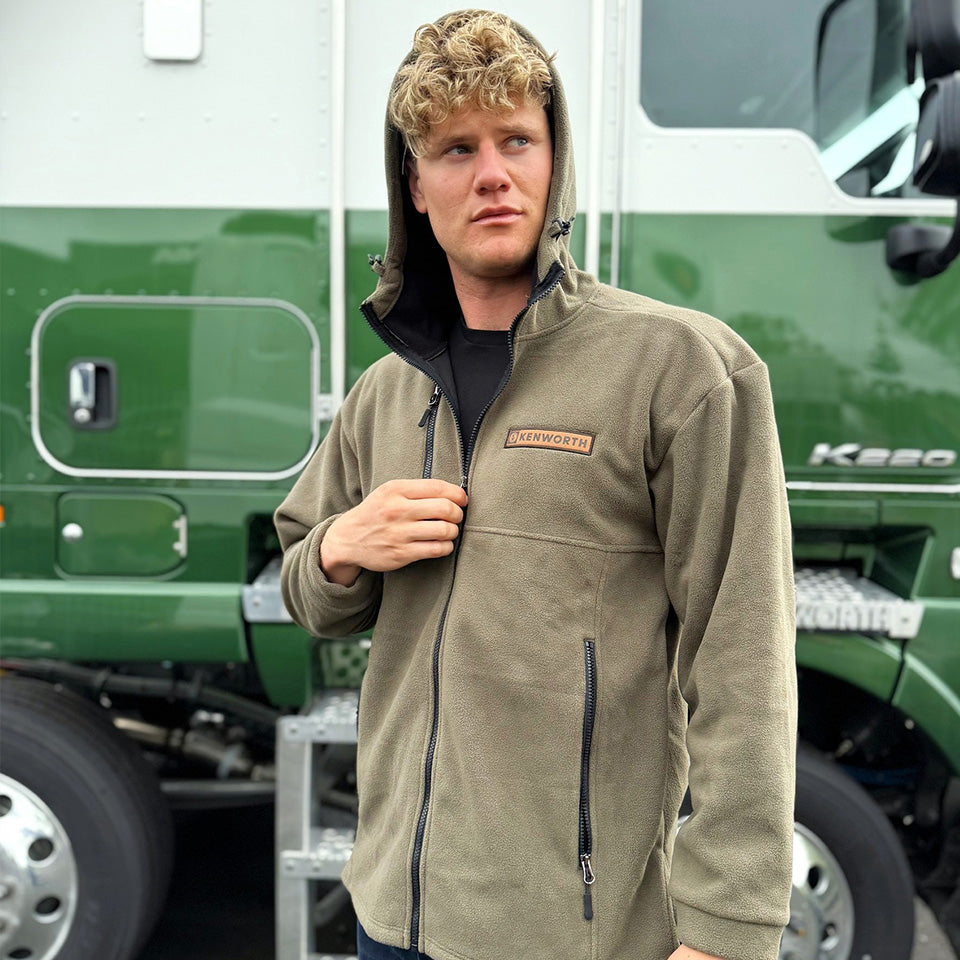 A man with curly blond hair wears an olive green FULL ZIP LONG SLEEVE BUSH SHIRT with a Kenworth patch, standing before a large green and white Kenworth K200 truck. He looks left, holding the shirt’s full zip.