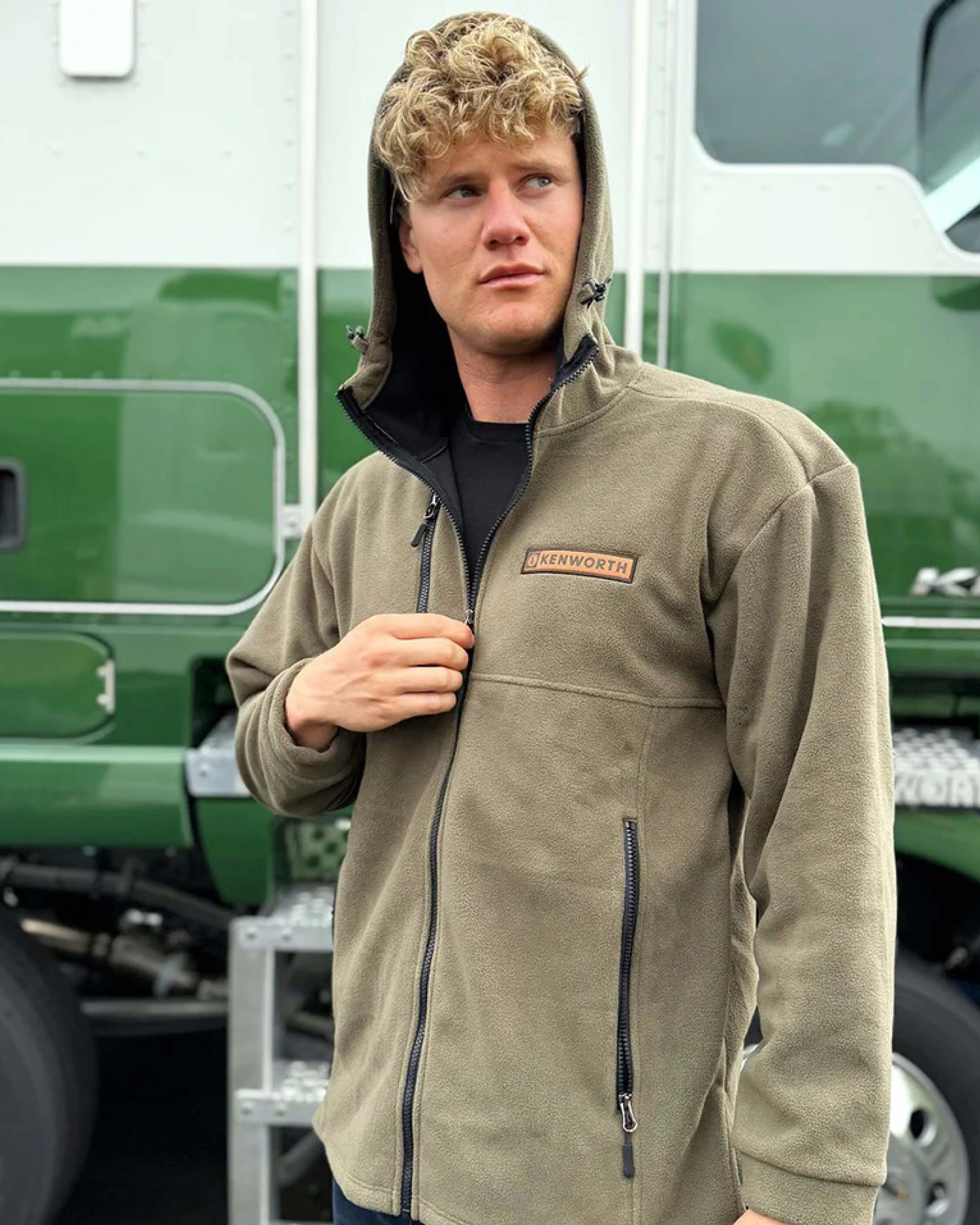 A man with curly blonde hair, wearing a khaki zip-up jacket with a Kenworth patch and the hood up, stands in front of a green and white truck, looking to his left.