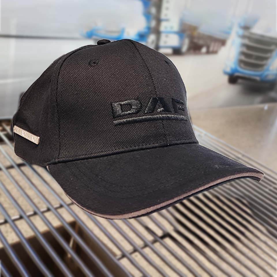 A black baseball cap with DAF embroidered on the front rests on a metal surface, with a blurred background showing blue trucks and an industrial setting.