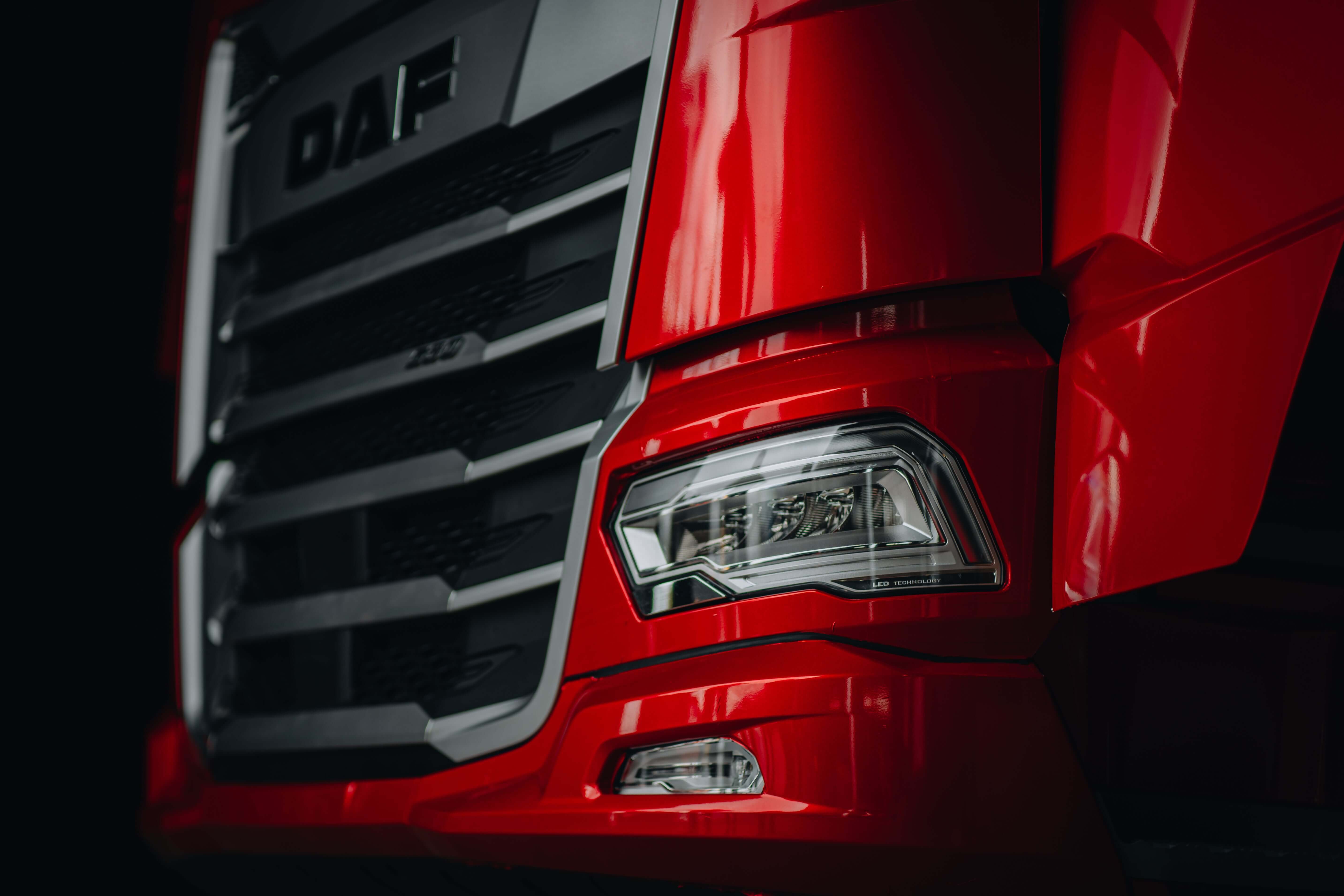 Close-up view of the front corner of a red DAF truck, showing the headlight, grille, and part of the bumper against a dark background.