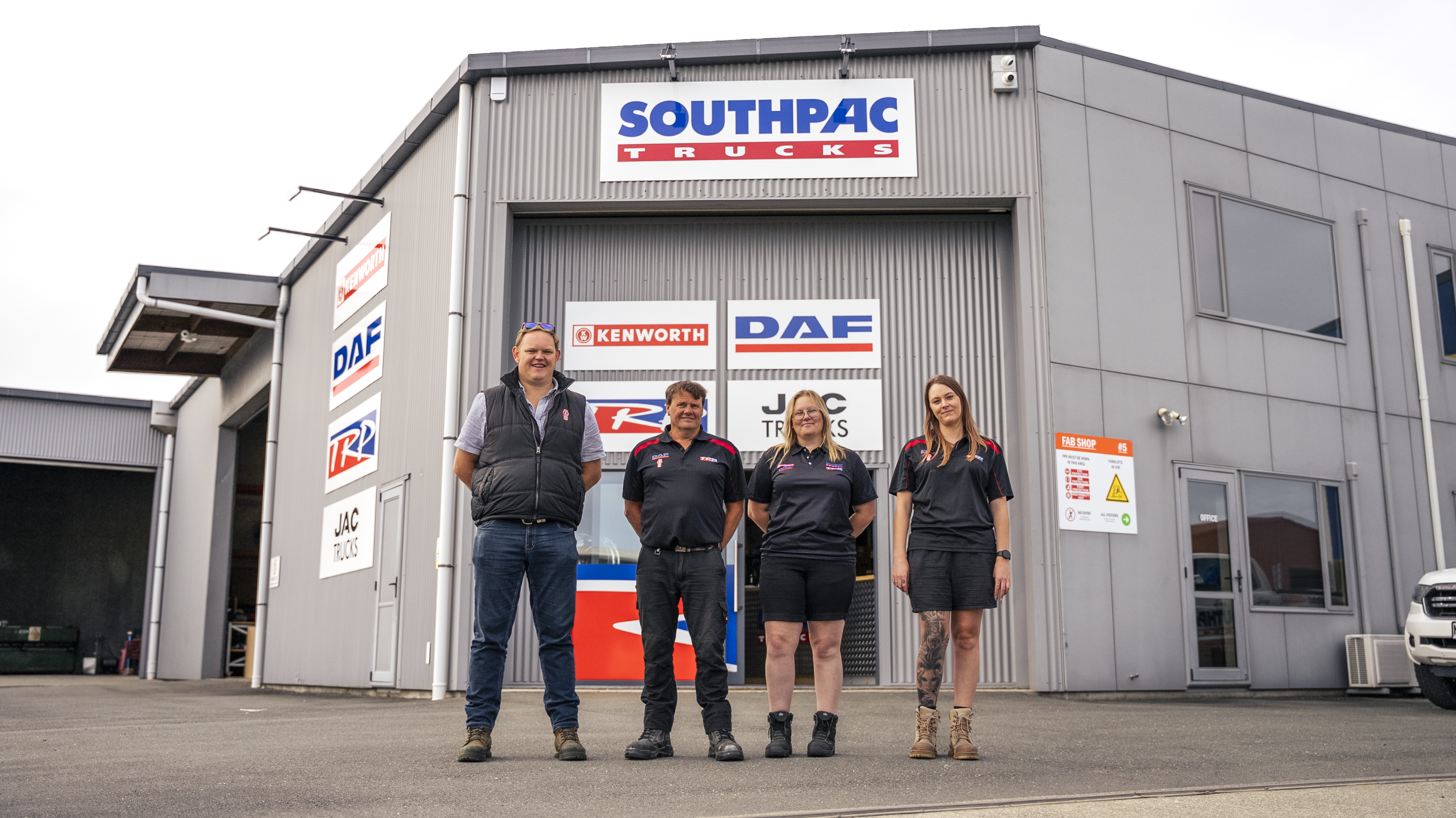Four people in uniforms stand in front of a Southpac Trucks building, which displays Kenworth and DAF signs. The group is smiling and posing for a photo outside the warehouse-style facility.