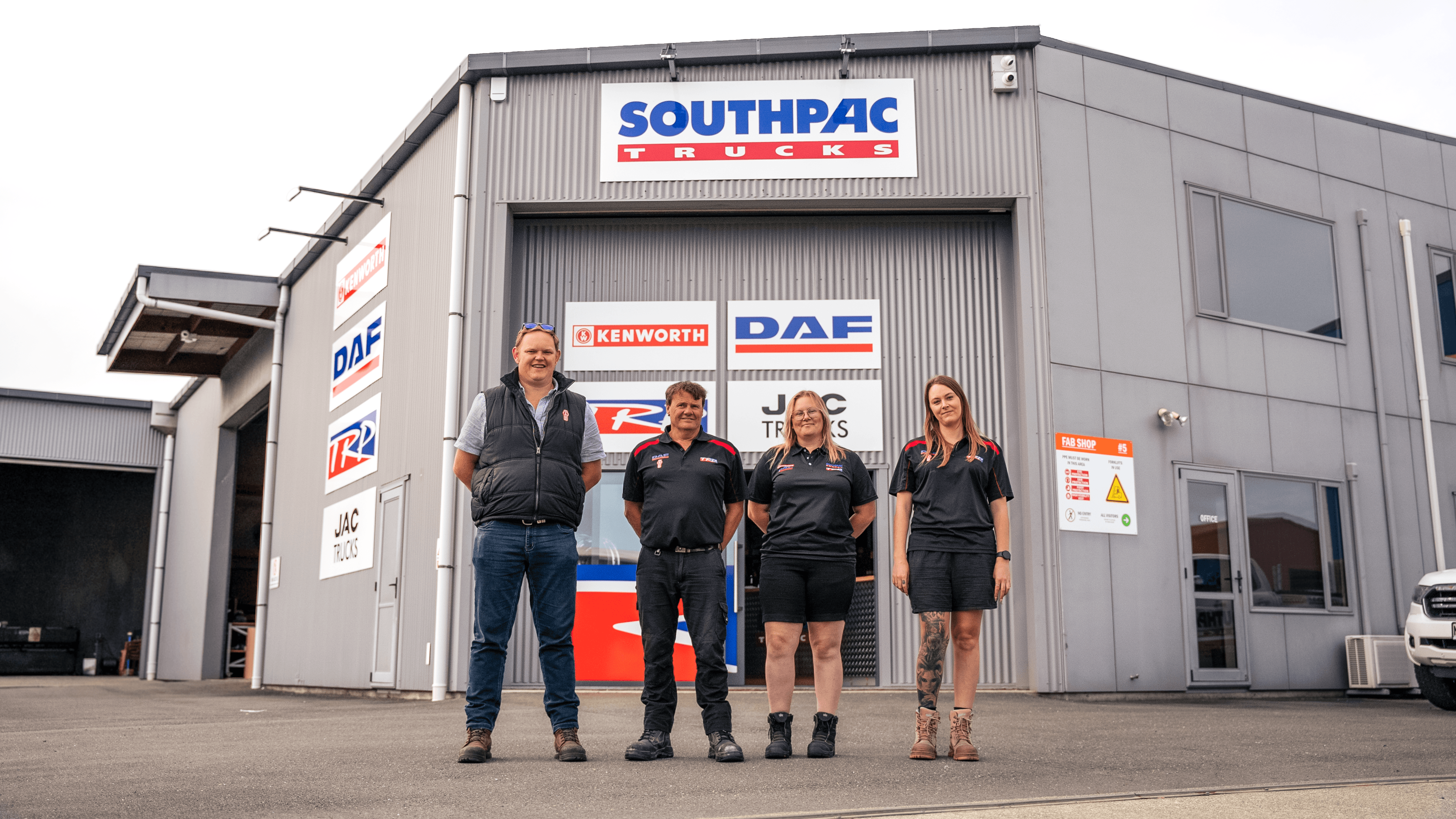 Four people in uniforms stand in front of a Southpac Trucks building, which displays Kenworth and DAF signs. The group is smiling and posing for a photo outside the warehouse-style facility.