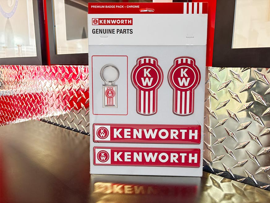 The DECAL KW SET CHROME PREMIUM displayed on a table includes a chrome Kenworth badge keychain, two round Kenworth decals, and two rectangular red logo badges. Silver diamond plate wall shown in the background.