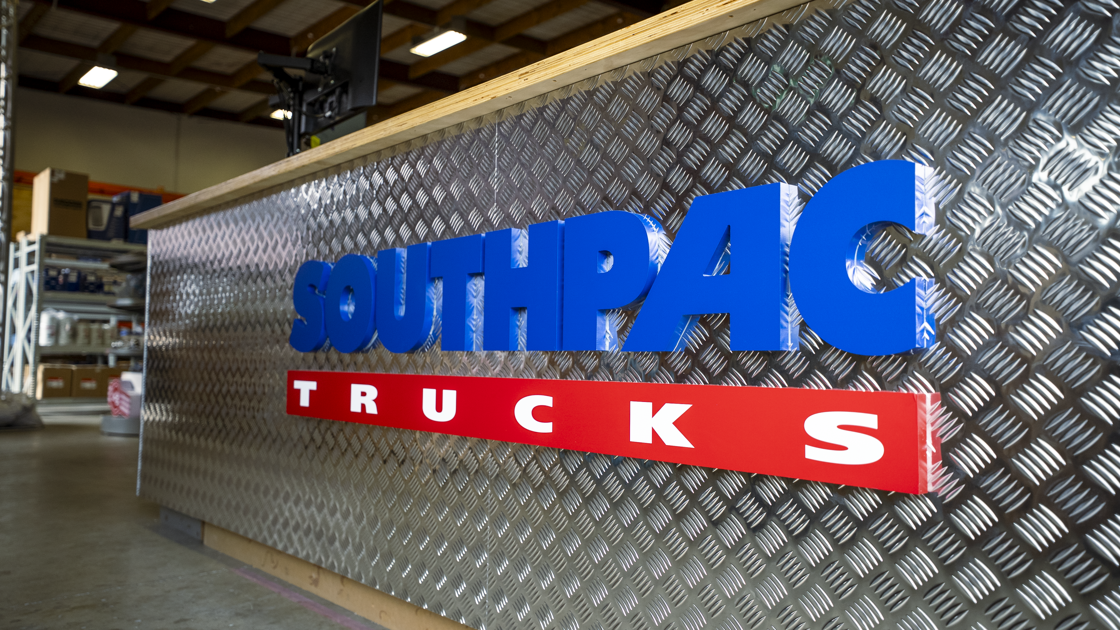 A metal counter with a diamond plate pattern displays bold blue letters spelling SOUTHPAC and red letters spelling TRUCKS inside a warehouse or industrial setting. Shelving and tools are visible in the background.