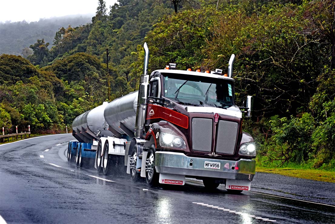 A large Kenworth tanker truck drives on a wet, winding road through a lush, green forested area on a rainy day, with trees and hills in the background.